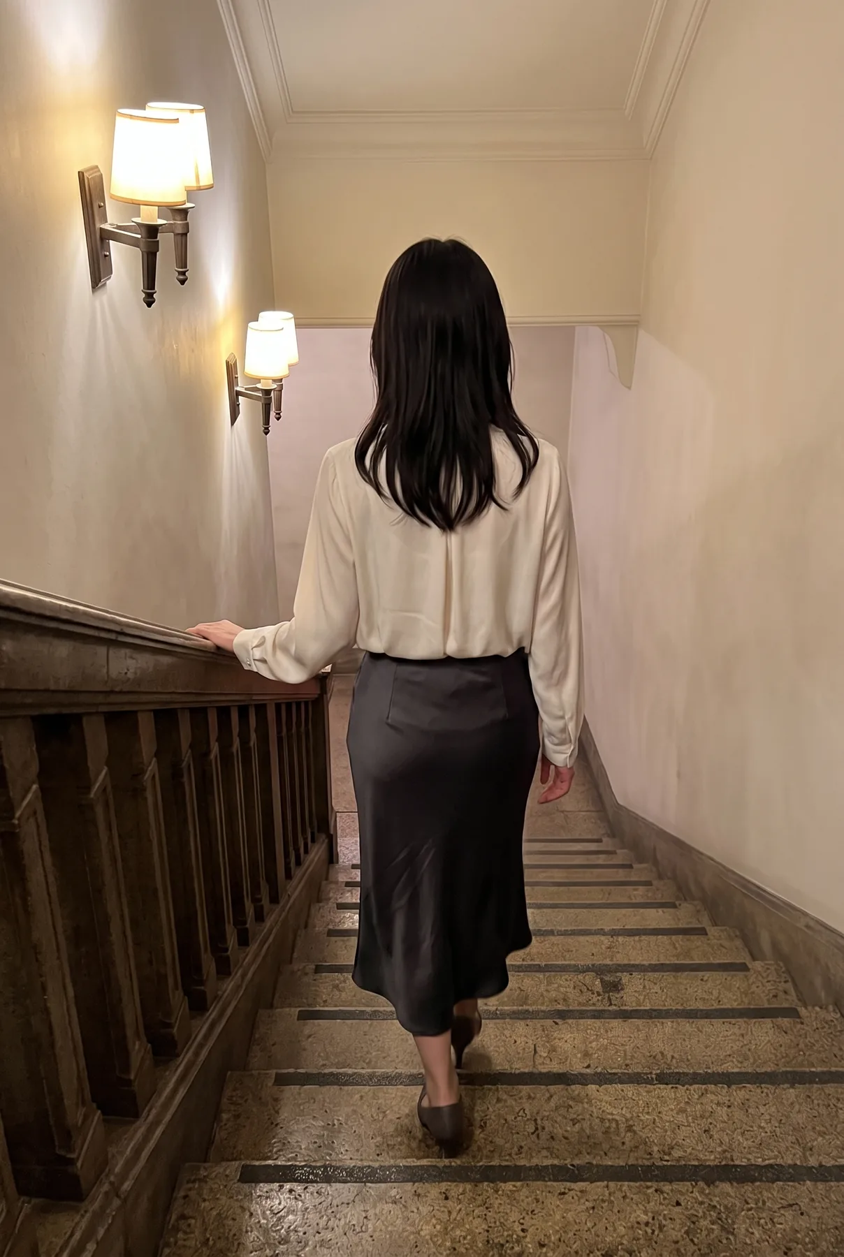 階段のある室内でクリーム色のブラウスと濃色のスカートを着て後ろ姿を見せる人物