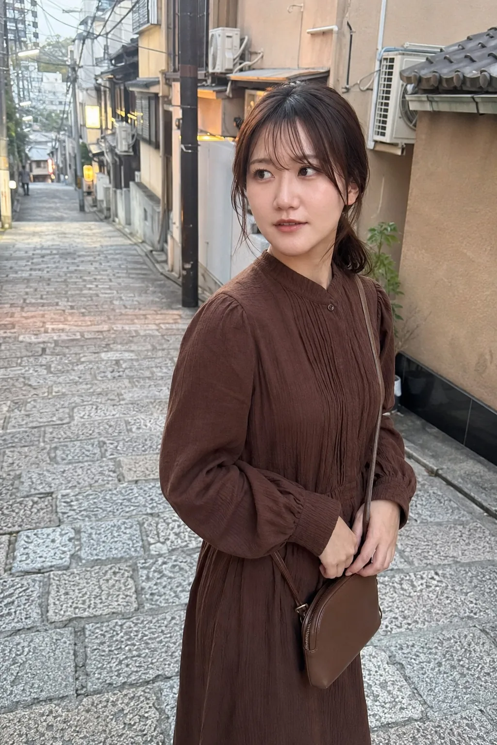 古い建物の並ぶ街路で茶色のワンピースを着てバッグを持ち横を見る人物