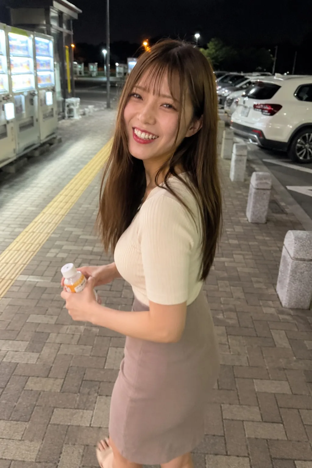 夜の駐車場でクリーム色の半袖トップスと淡い色のスカートを着て振り向き笑う人物