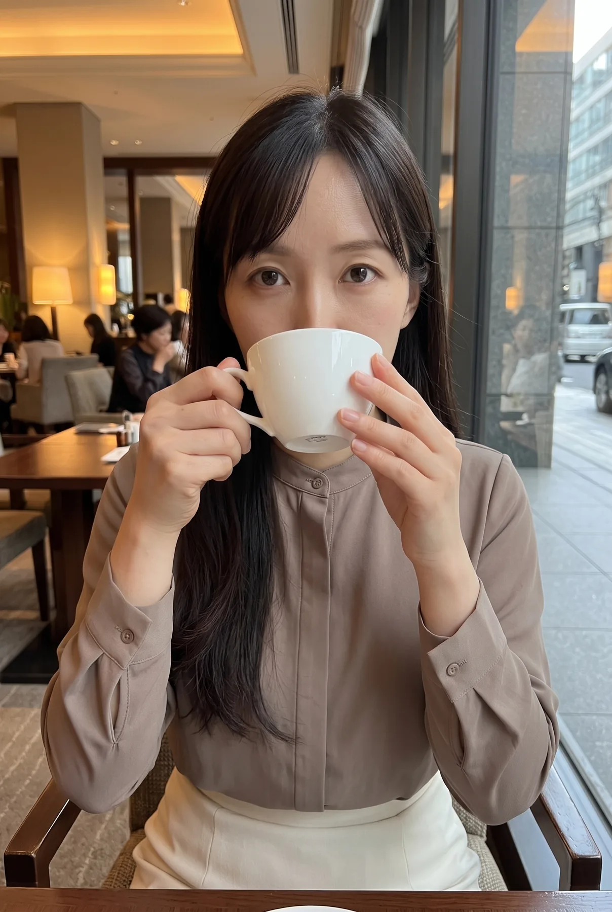 カフェの席で淡い茶色のブラウスと白いスカートを着て白いカップを口元に持つ人物