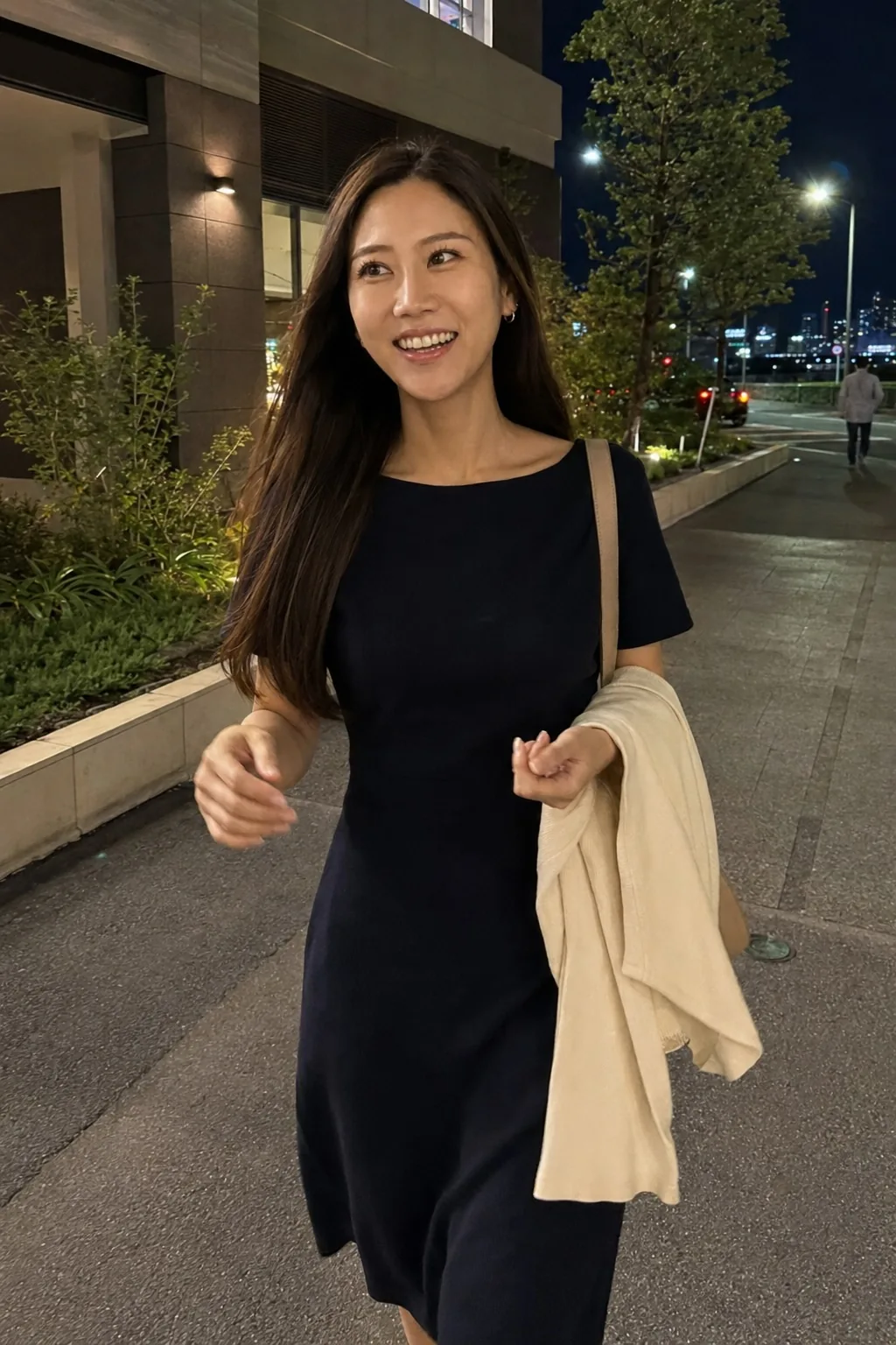 夜の街路で紺のワンピースを着て白いカーディガンを腕に掛けて笑う人物