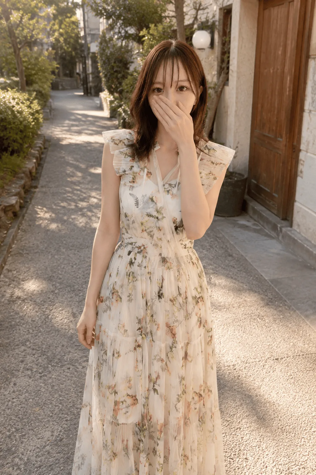 夕方の街路で花柄のワンピースを着て立ち、片手を口元に当てて正面を見る人物の3/4身
