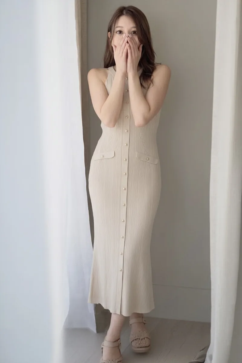 窓辺でベージュのノースリーブのニットワンピースを着て、両手で鼻と口元を覆い、正面を見る女性の全身
