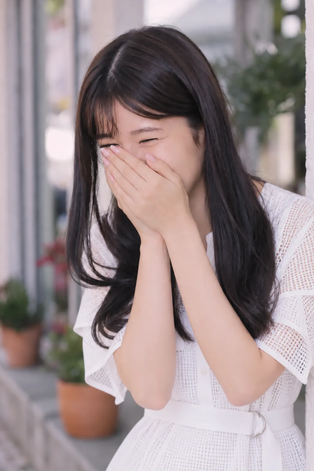白いワンピースの女性が両手で鼻と口元を覆い、目を細めて大きく笑う。顔と上半身を近くから捉えた縦構図。